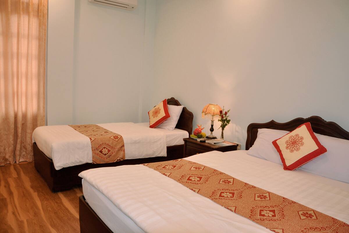 A hotel room with two beds, each with a patterned bedspread and pillows, a nightstand with a lamp and a vase of flowers, and a wooden floor.