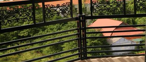 Ein Balkon mit schwarzem Metallgeländer und Blick auf ein Gebäude mit rotem Dach und Grünpflanzen im Hintergrund.