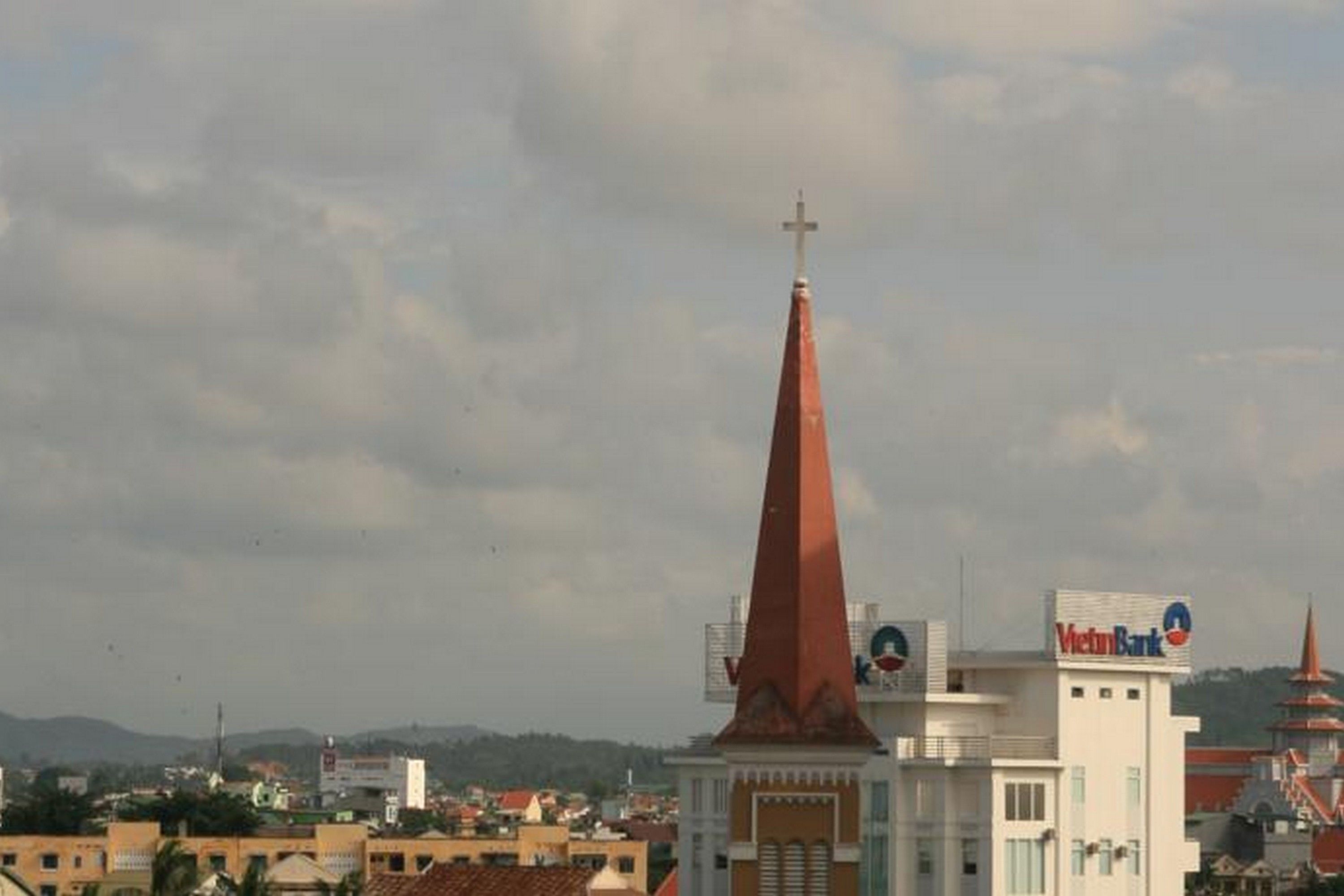 A church with a tall red spire and a cross at the top, a building with a sign for VietinBank, and a mountainous background.