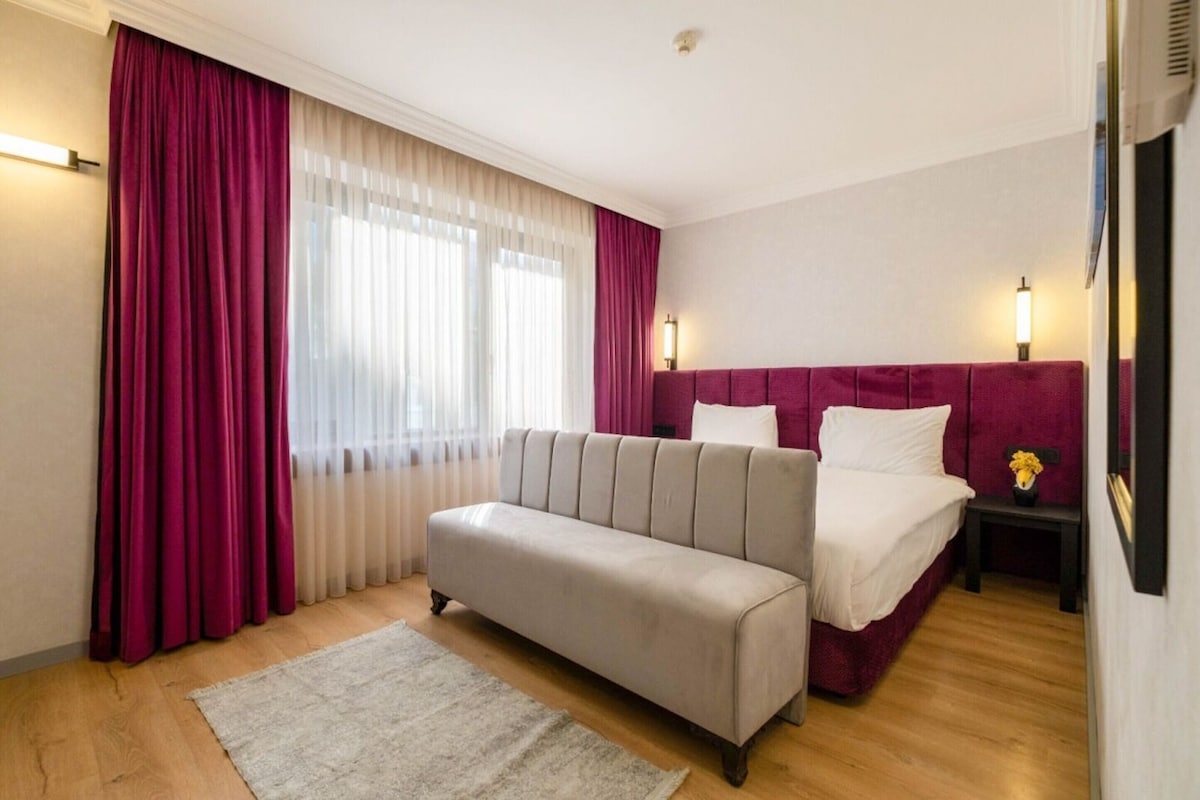 Ein Hotelzimmer mit Bett, Sofa, Nachttisch, Lampe und einem Fenster mit Vorhängen.