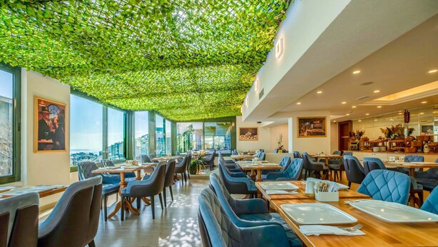 En restaurang med högt i tak, en grönskande takdekoration, modern möblering och stora fönster med utsikt över staden.