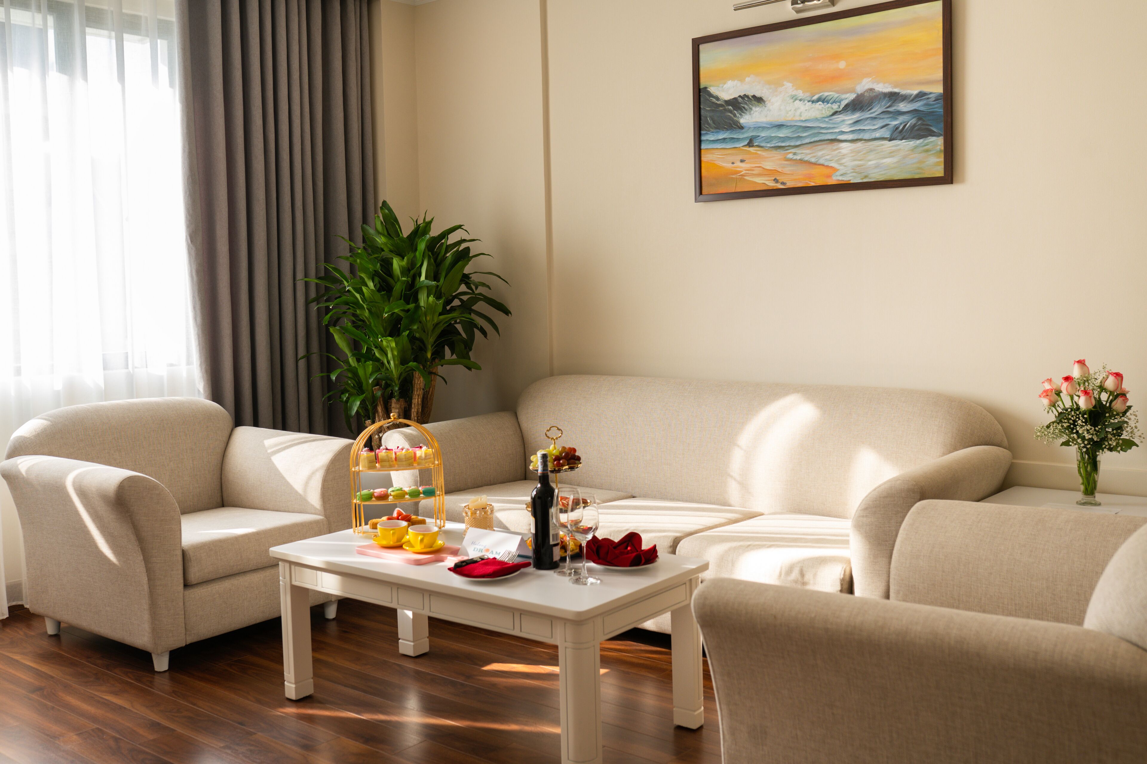 A living room with a beige sofa, a white coffee table with food and drinks, a potted plant, and a painting of a beach scene.