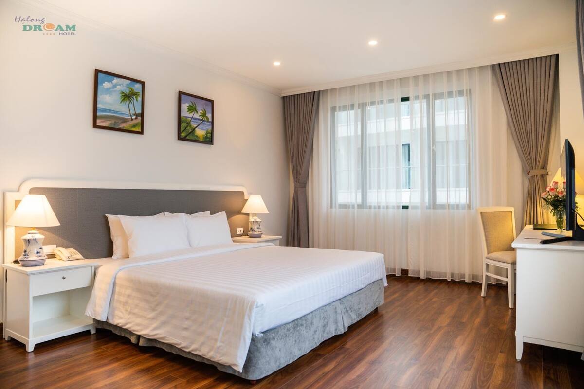 A hotel room with a large bed, two bedside tables with lamps, a desk with a chair, and a window with curtains.