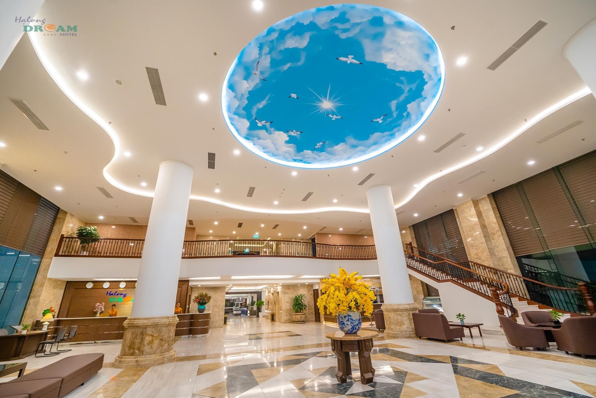 A spacious lobby with a large, illuminated ceiling mural of a sky with clouds and birds, a reception desk, a staircase, and a seating area with a vase of yellow flowers.