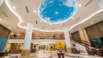 A spacious lobby with a large, illuminated ceiling mural of a sky with clouds and birds, a reception desk, a staircase, and a seating area with a vase of yellow flowers.