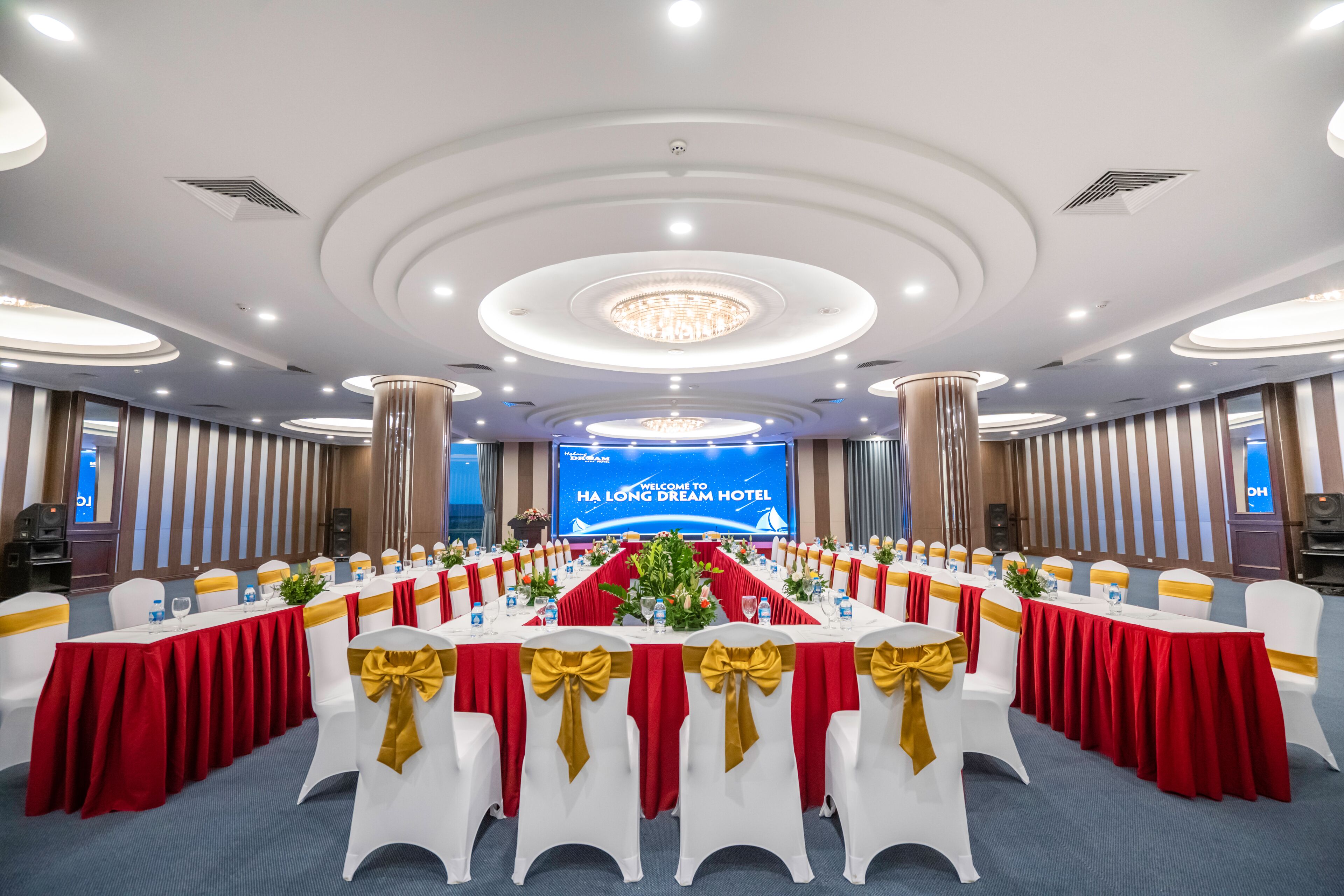 Een vergaderruimte met ronde tafels, witte stoelen met gouden linten, rode tafelkleden en een groot scherm waarop staat: 'Welcome to HA Long Dream Hotel'.