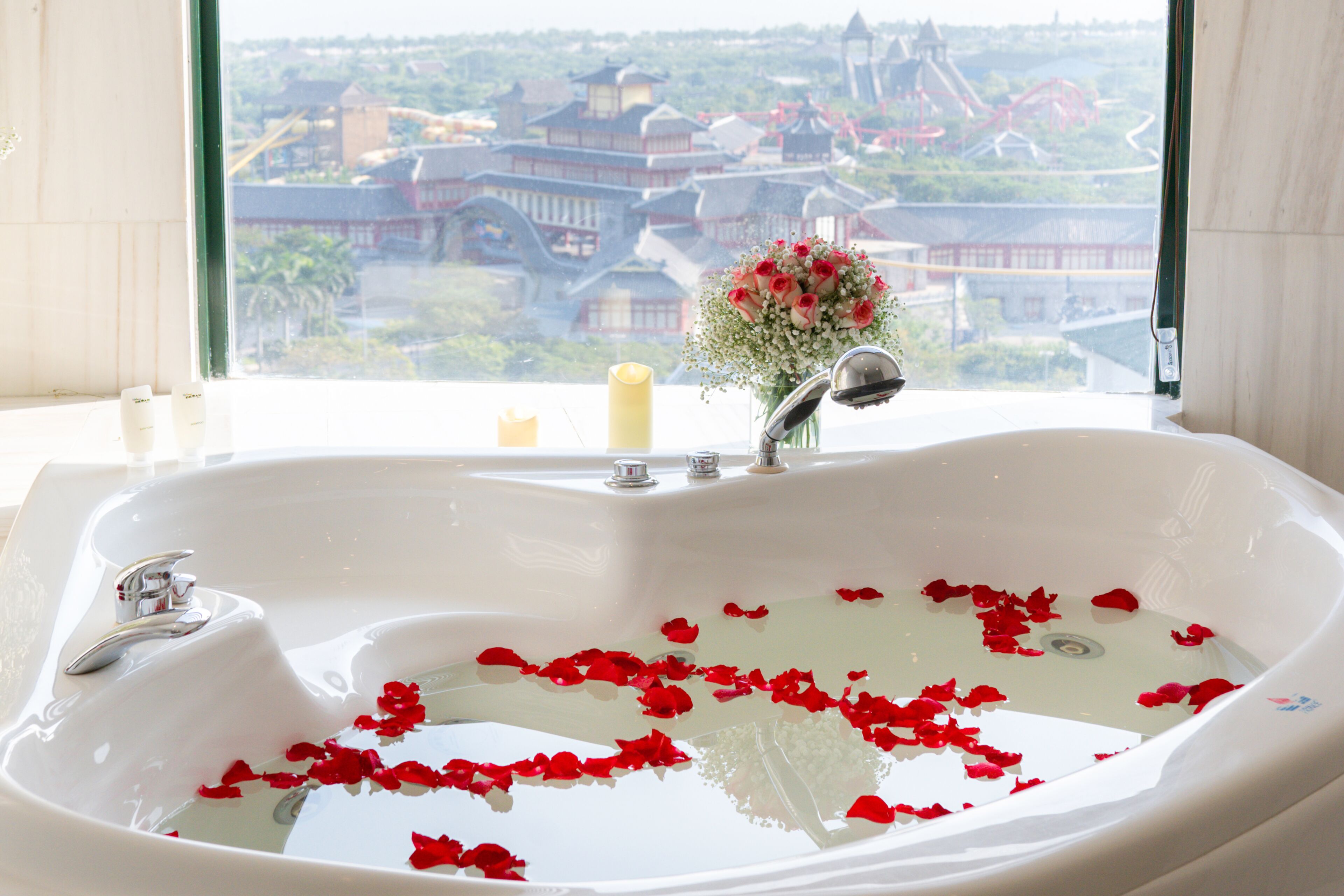 A bathroom with a large bathtub filled with water and rose petals, a window with a view of traditional buildings, and a vase with flowers on the windowsill.