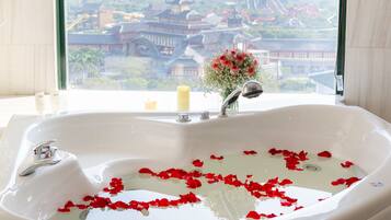 A bathroom with a large bathtub filled with water and rose petals, a window with a view of traditional buildings, and a vase with flowers on the windowsill.