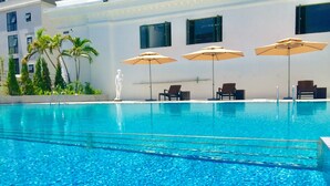 En swimmingpool med klart, blåt vand, omgivet af liggestole og parasoller, samt en statue i baggrunden.