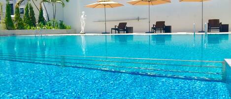 A swimming pool with clear blue water, surrounded by lounge chairs and umbrellas, and a statue in the background.