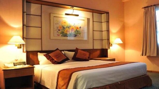 Ein Hotelzimmer mit einem großen Bett, zwei Nachttischen, einem Tischchen, einem Bild an der Wand und einem Fenster mit Vorhängen.