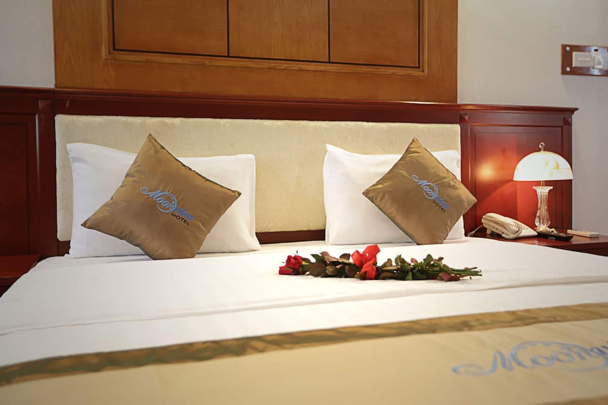 A hotel room with a bed, two pillows with the hotel logo, a bedside table with a lamp, and a bouquet of flowers.