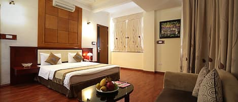 Habitación de hotel con una cama grande, cabecera de madera, mesita de noche con lámpara, pequeña mesa con fruta, un sofá y cortinas.