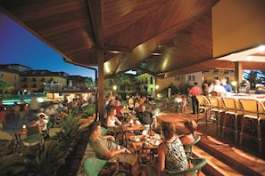 Guests can enjoy meals in a lively outdoor dining area with comfortable seating and a pool in the background.