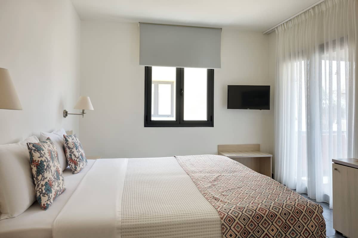 Une chambre d’hôtel avec un lit, un téléviseur fixé au mur, une fenêtre avec un store et une table de chevet.