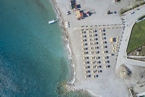 Une vue aérienne d’une plage, avec des rangées de parasols et de chaises de plage, une jetée qui s’avance dans l’eau et un parking où sont stationnés des véhicules.