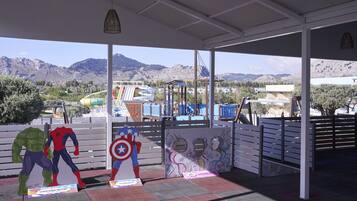 A covered outdoor area with a view of mountains, a playground with slides, and superhero cutouts.