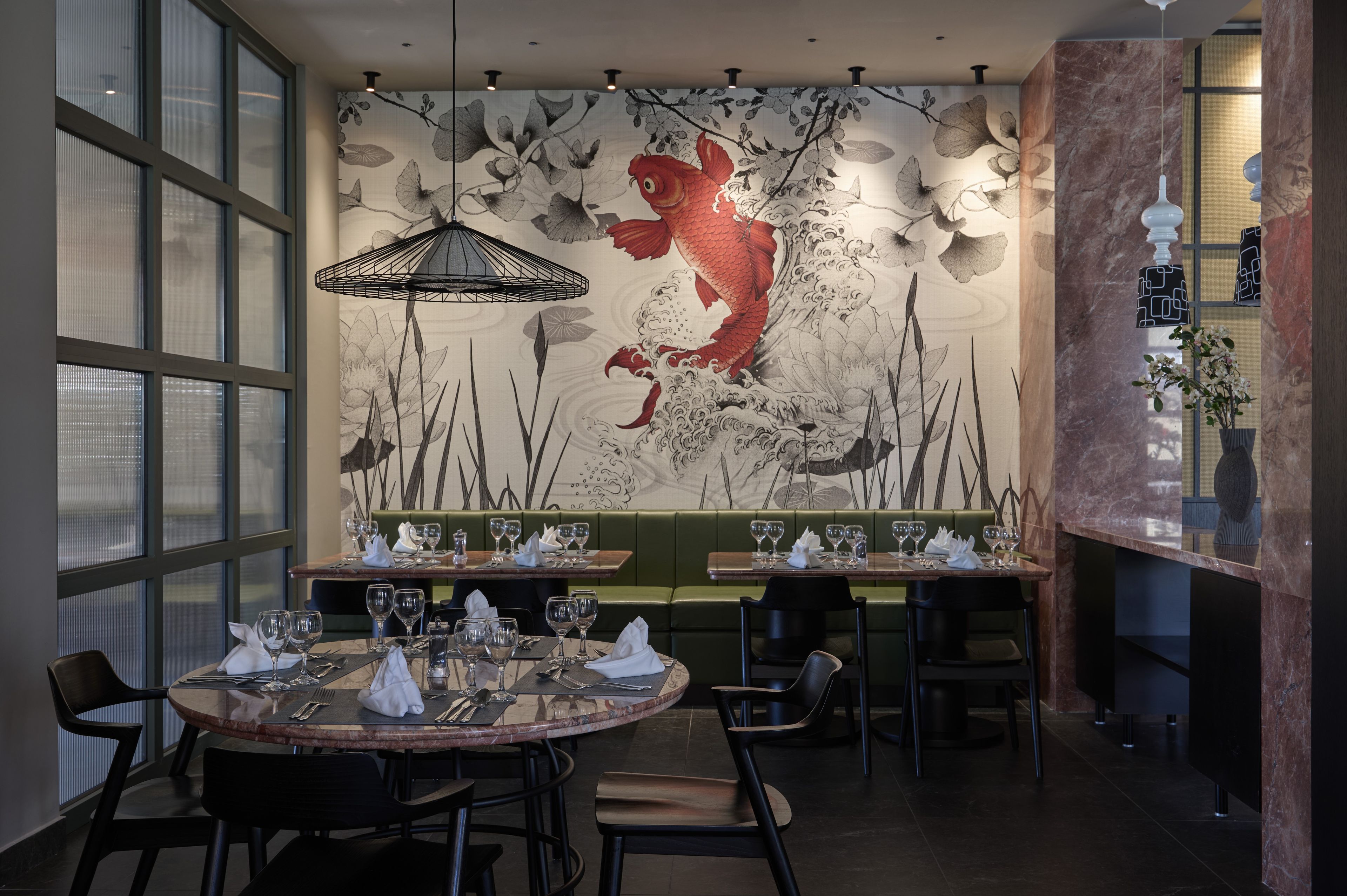 A restaurant interior with a mural of a koi fish and lotus flowers, dining tables set with glasses and cutlery, and a green booth seating area.