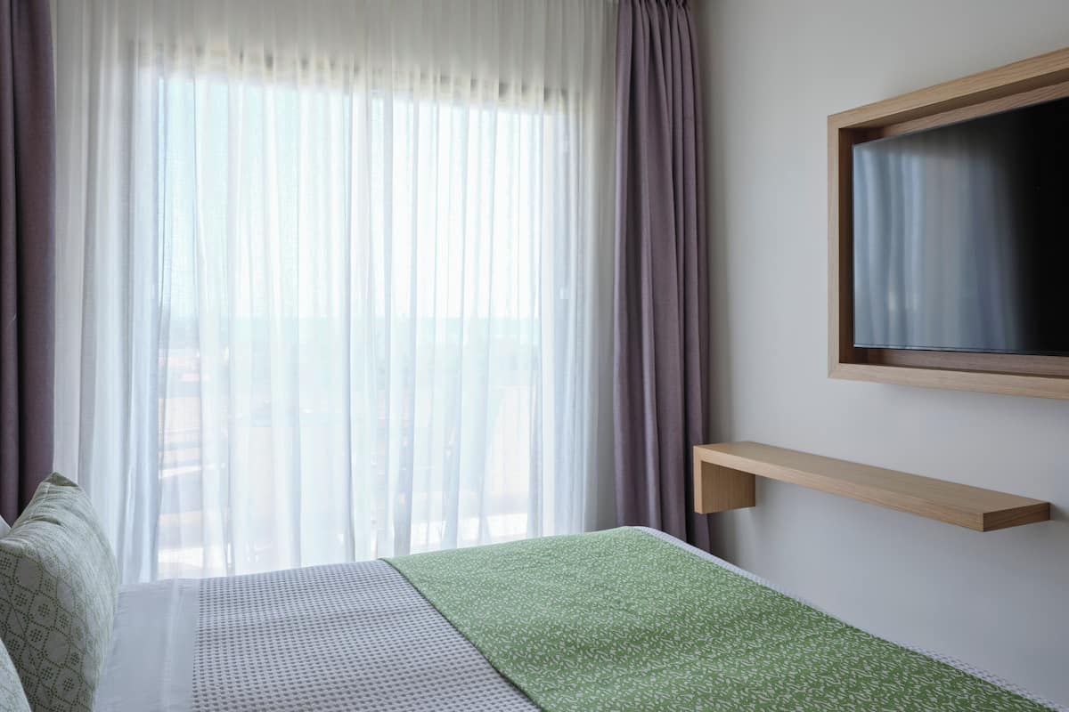 Une chambre d’hôtel avec un lit, un téléviseur fixé au mur et une fenêtre avec des rideaux légers.