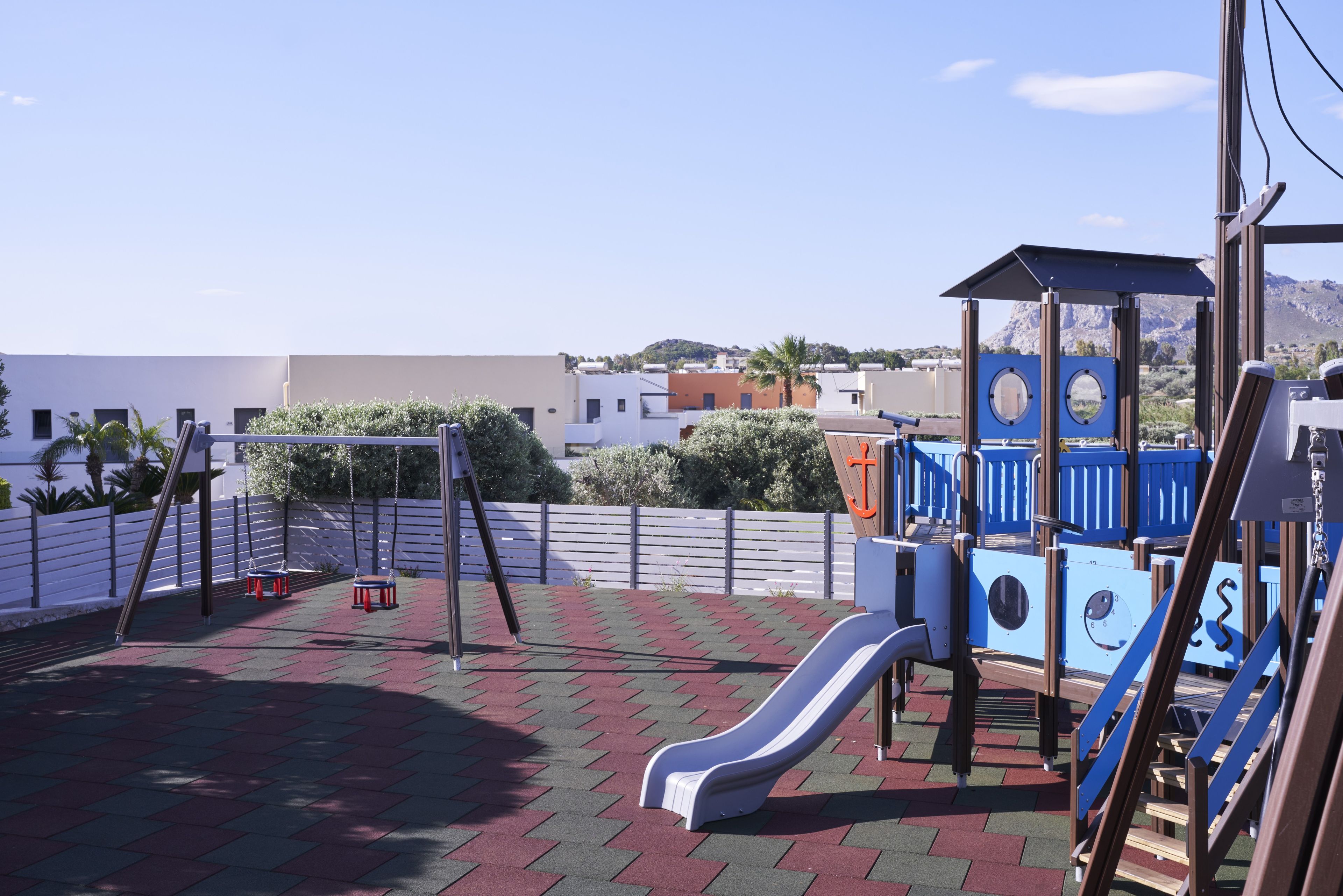 A playground with a slide and swings, surrounded by a white fence and a view of buildings and hills.
