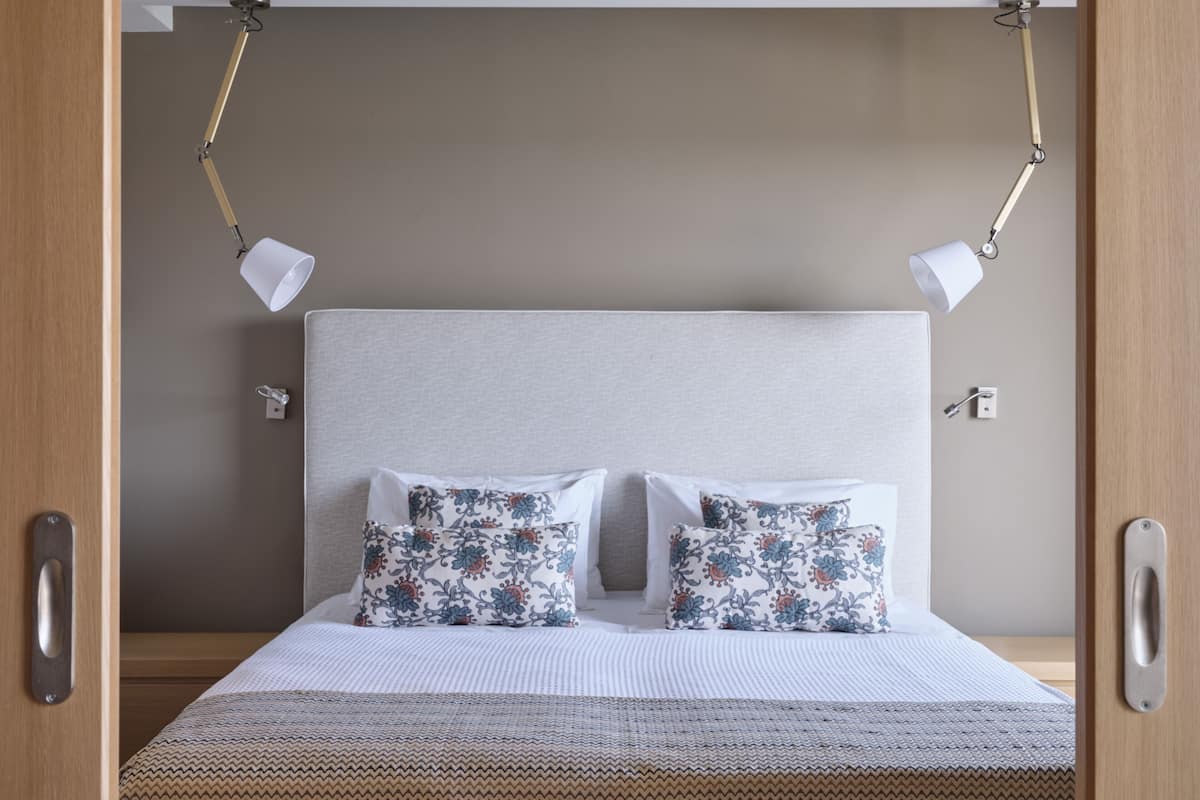 Un lit bien fait, agrémenté de deux coussins à motifs floraux, d’une tête de lit sobre et de deux lampes de chevet.