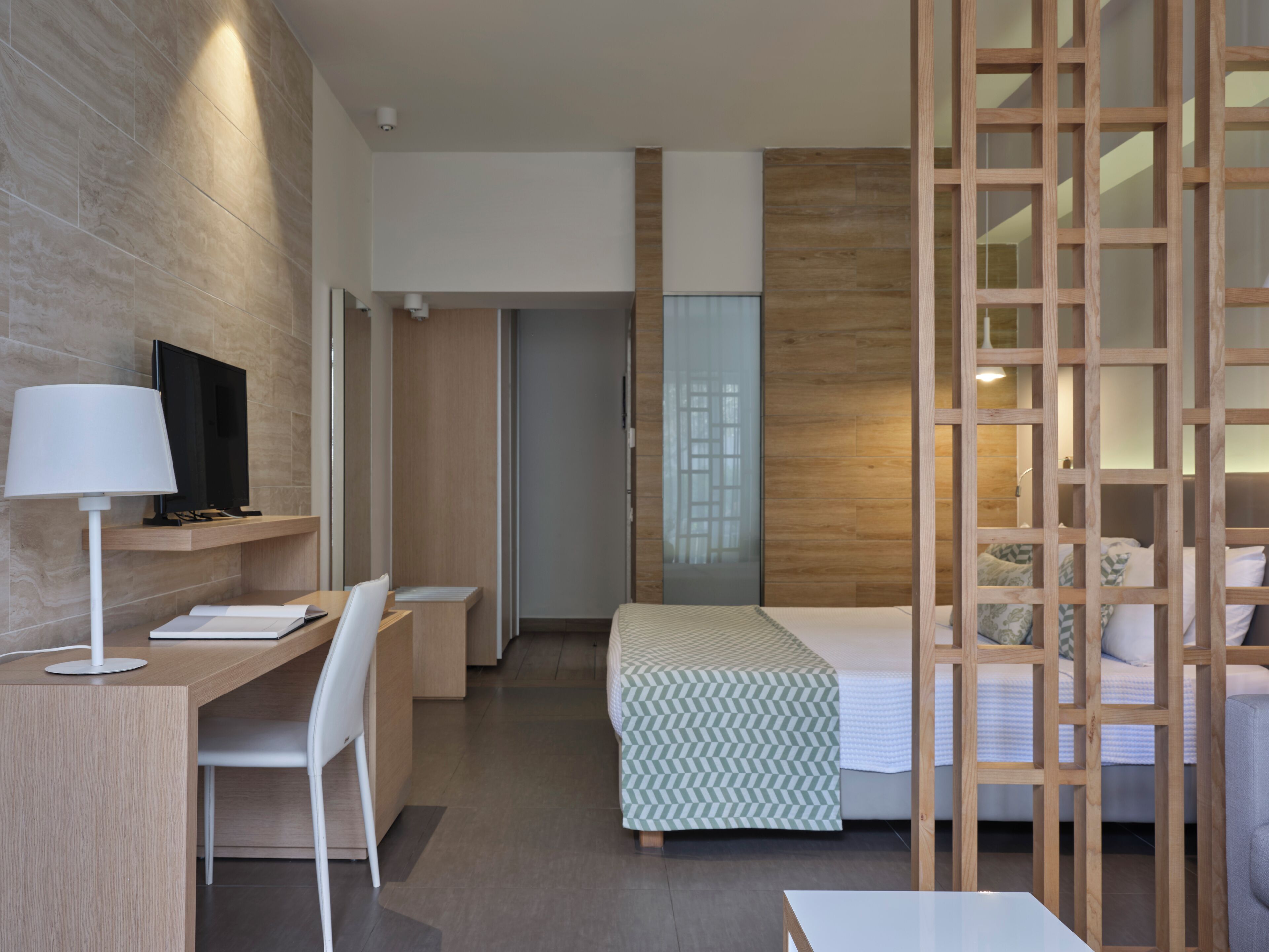 Una habitación de hotel moderna con un escritorio de madera, una cama con colcha estampada, un televisor en la pared y una partición de madera.