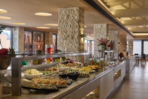 Buffet variado com saladas, sanduíches e aperitivos, além de um pilar de pedra e uma vitrine de vidro.