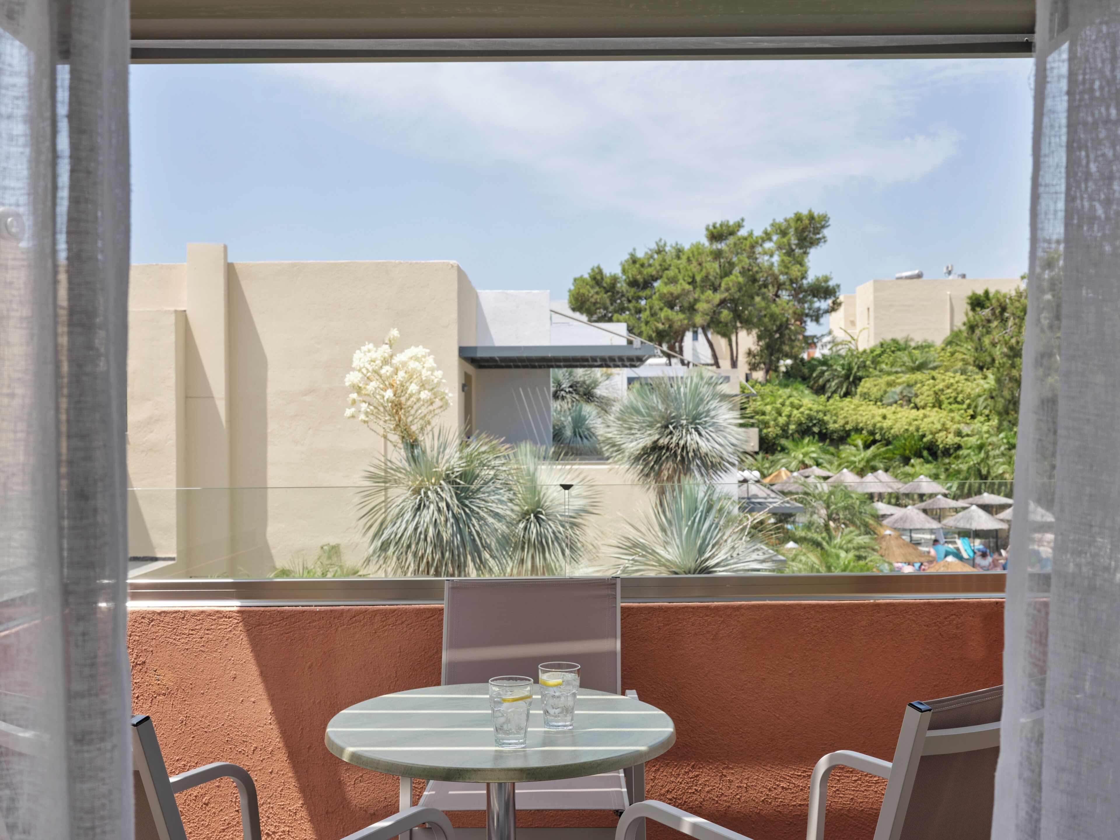Un balcón con mesa y sillas, con vista a una zona de piscina con sombrillas y palmeras.