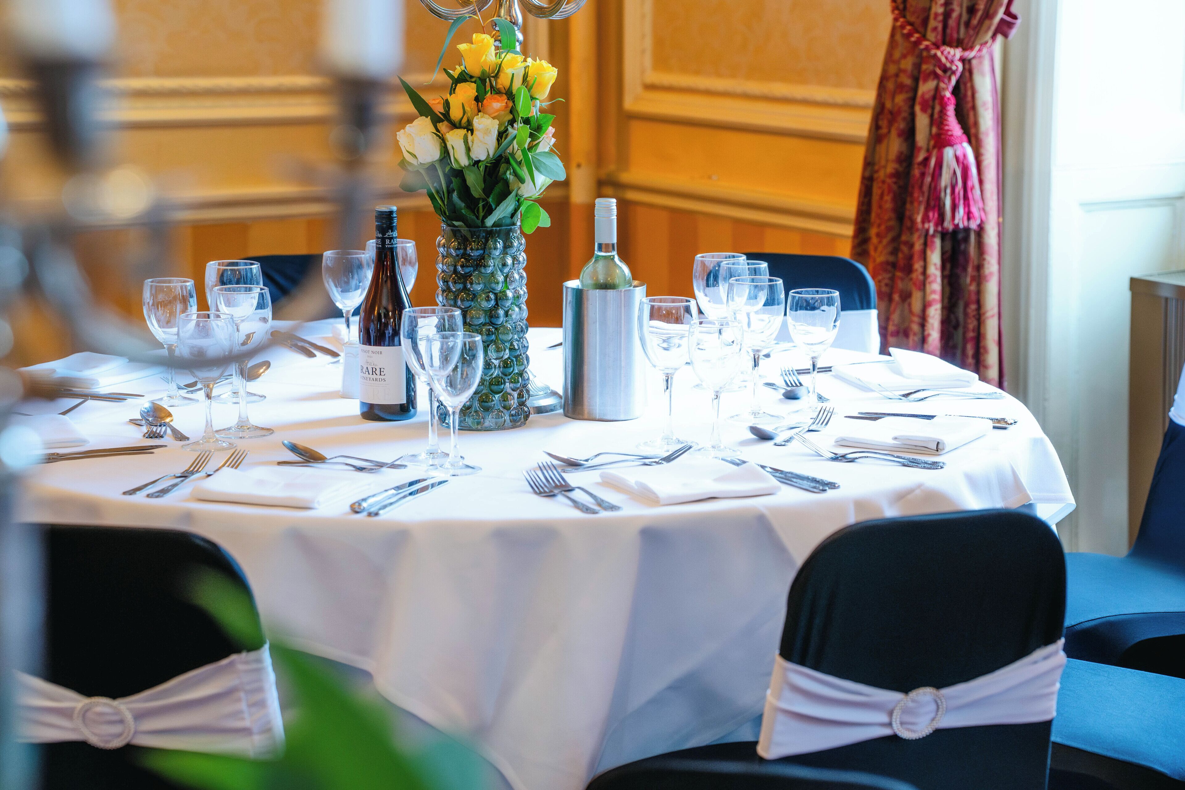 A round dining table set with white tablecloth, wine glasses, and a vase with flowers.