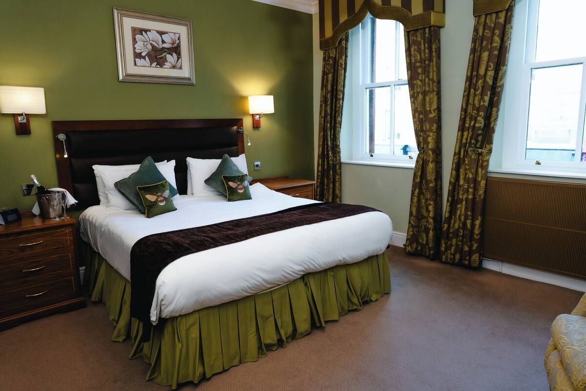 A neatly made bed with a headboard, two bedside tables with lamps, a framed picture on the wall, and a window with curtains.