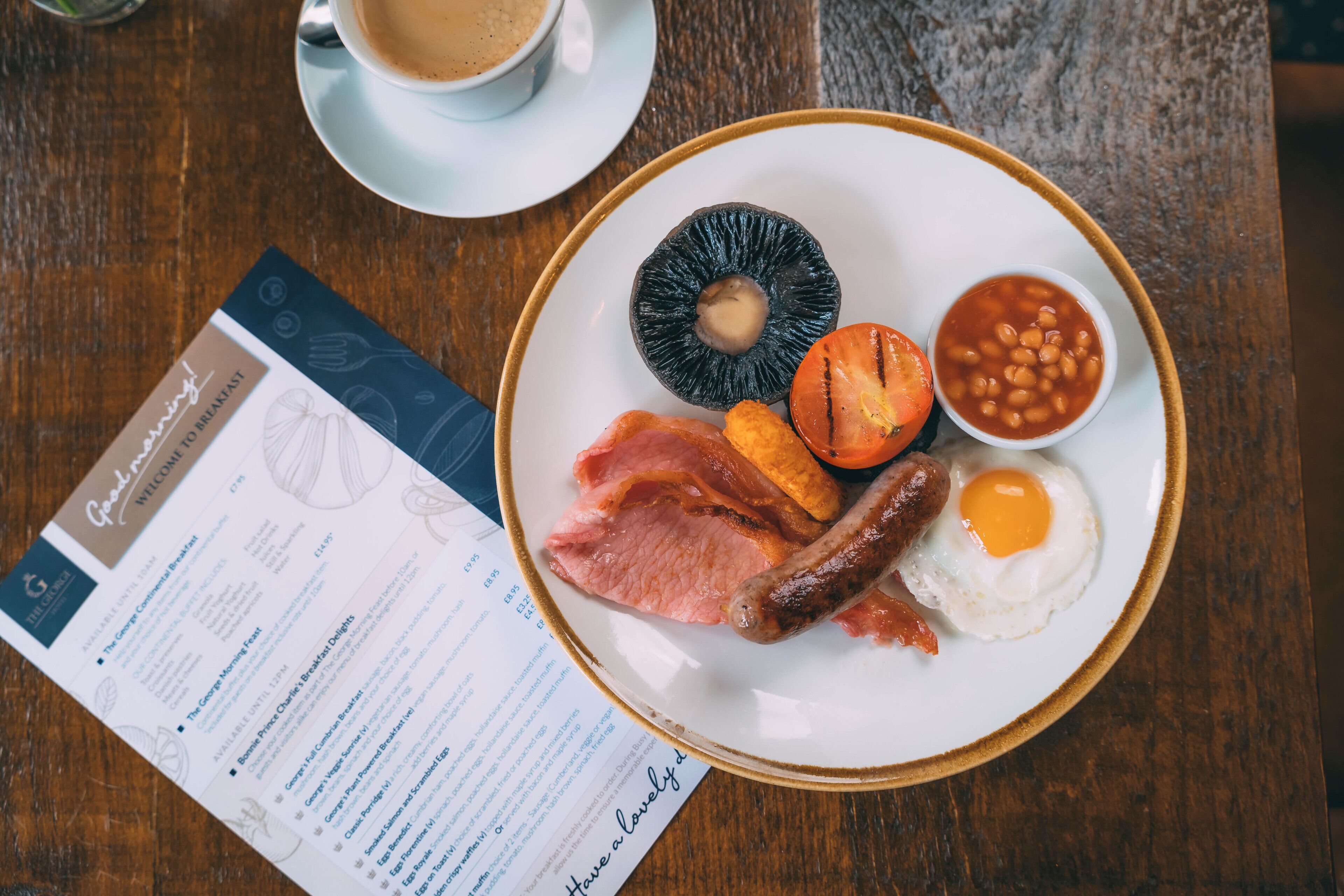 A breakfast plate with eggs, bacon, beans, and a tomato on a wooden table.