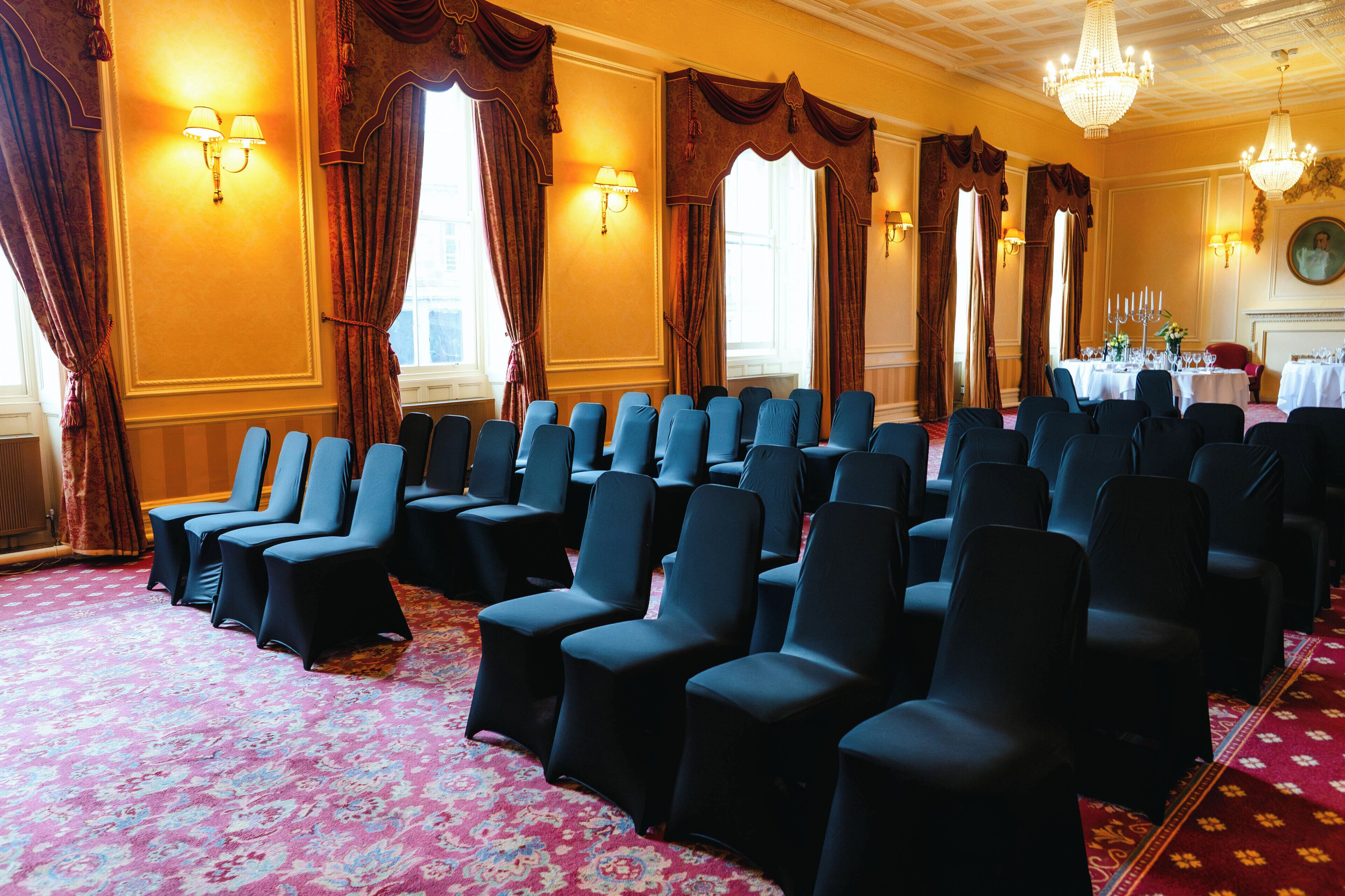 A conference room with rows of chairs, large windows with curtains, chandeliers, and a carpeted floor.