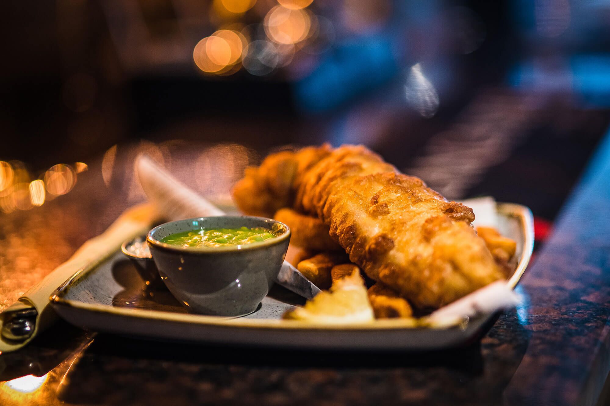 Una bandeja con un gran trozo de pescado frito, un bol de salsa verde y papas fritas.