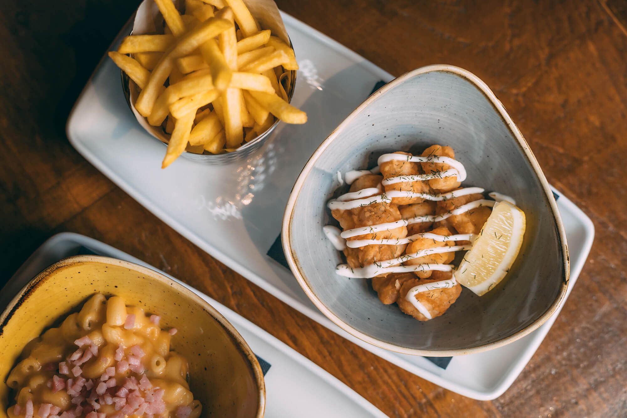 A tray with three dishes: French fries, a bowl of breaded and fried items with white sauce and lemon, and a bowl of macaroni with cheese and ham.
