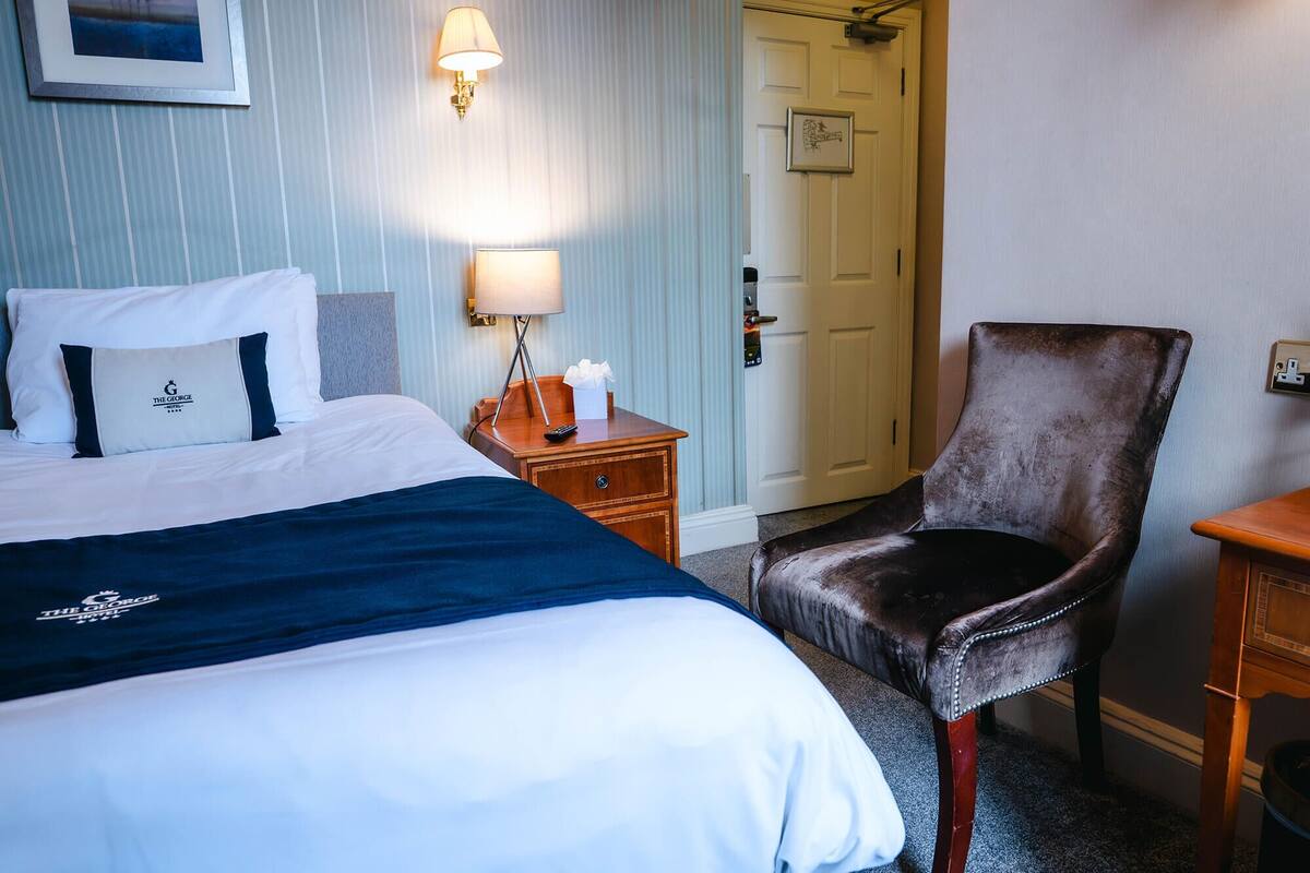 A hotel room with a bed, a chair, a nightstand, a lamp, and a door.