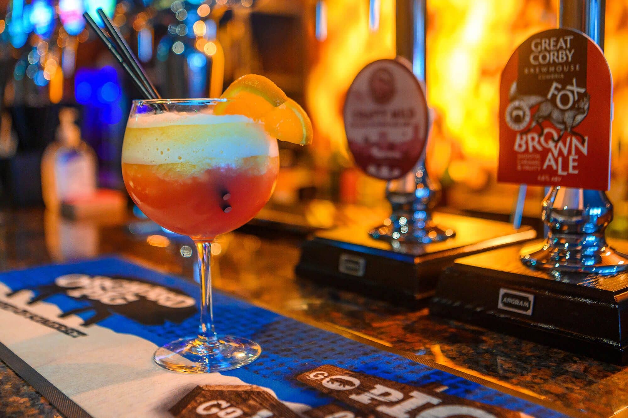 A cocktail with a layered drink and an orange garnish, a beer tap labeled "Great Corby," and a coaster with "Go Pro" text.