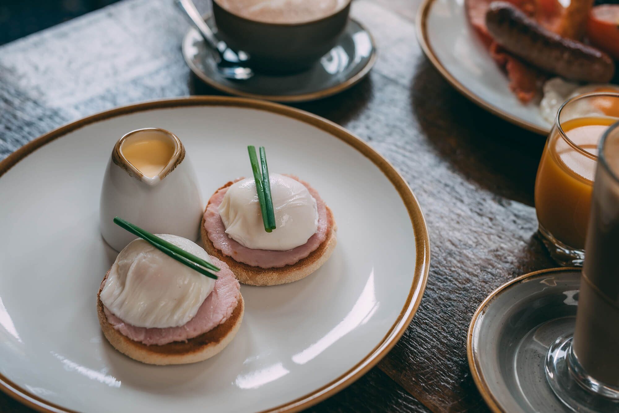 Una mesa de madera con dos sándwiches huevos Benedict, una taza de café y un vaso de jugo de naranja.