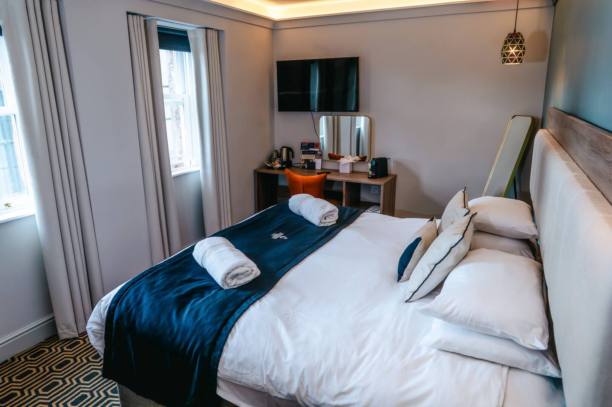 A hotel room with a neatly made bed, a desk with a computer and a lamp, a TV mounted on the wall, and a window with curtains.