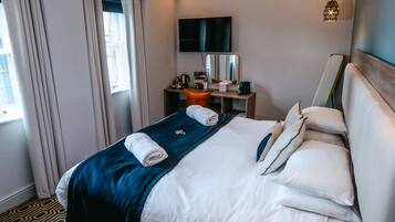 A hotel room with a neatly made bed, a desk with a computer and a lamp, a TV mounted on the wall, and a window with curtains.