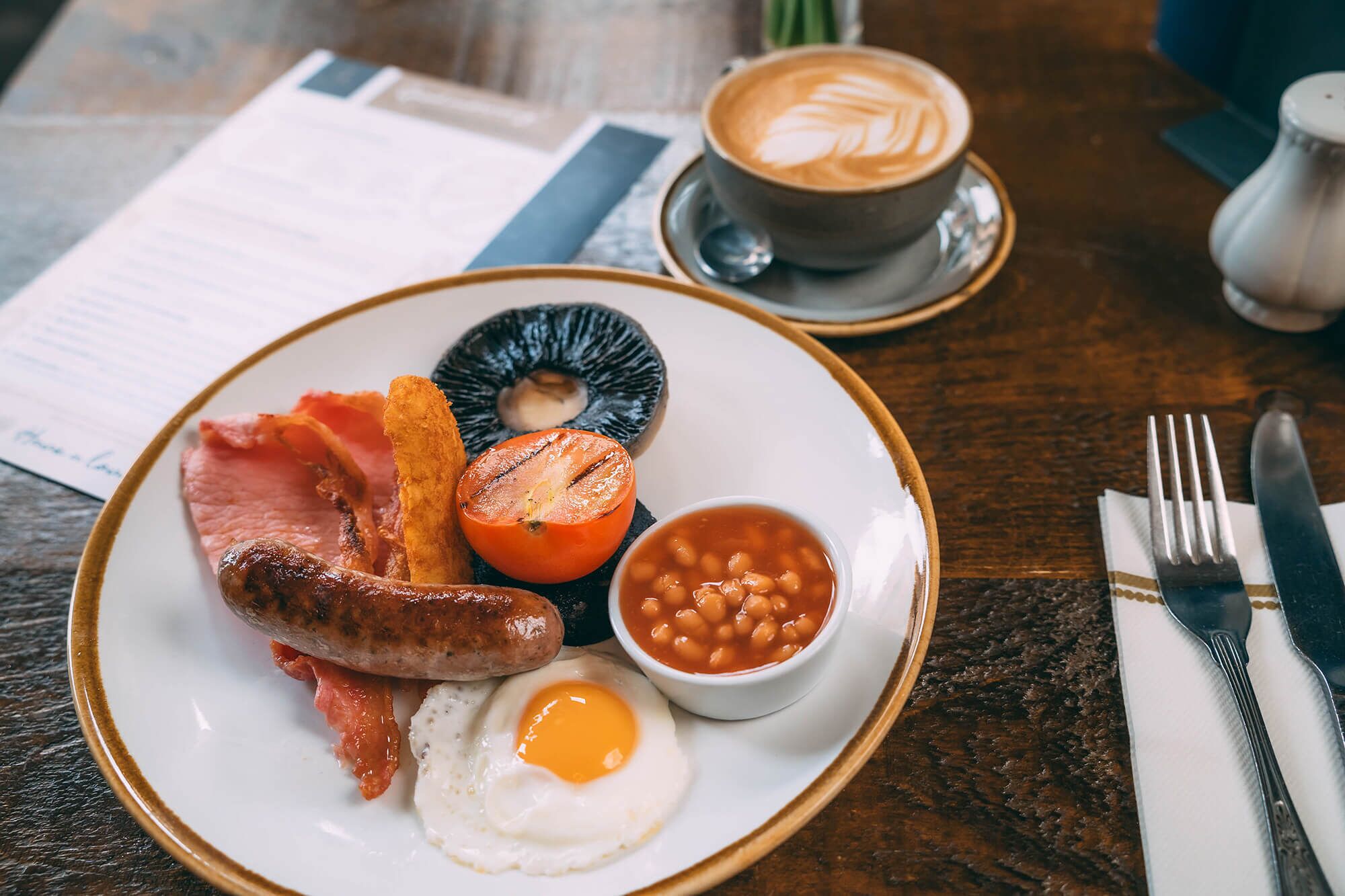 A breakfast plate with bacon, sausage, egg, baked potato, baked beans, and a slice of tomato on a wooden table.
