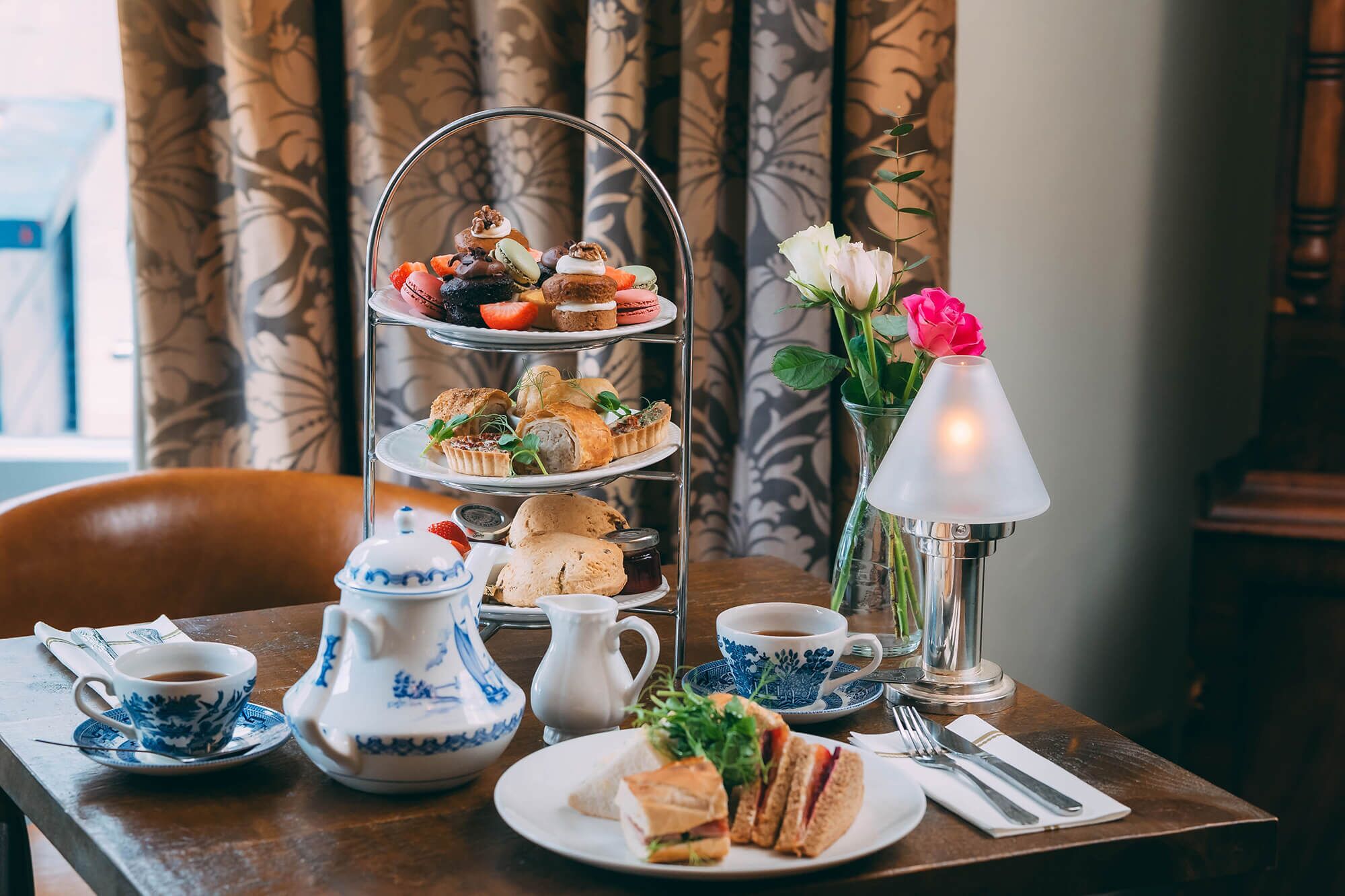 A tiered dessert stand with assorted pastries, a floral vase with a pink rose, a teapot, cups, and a table set for a meal.