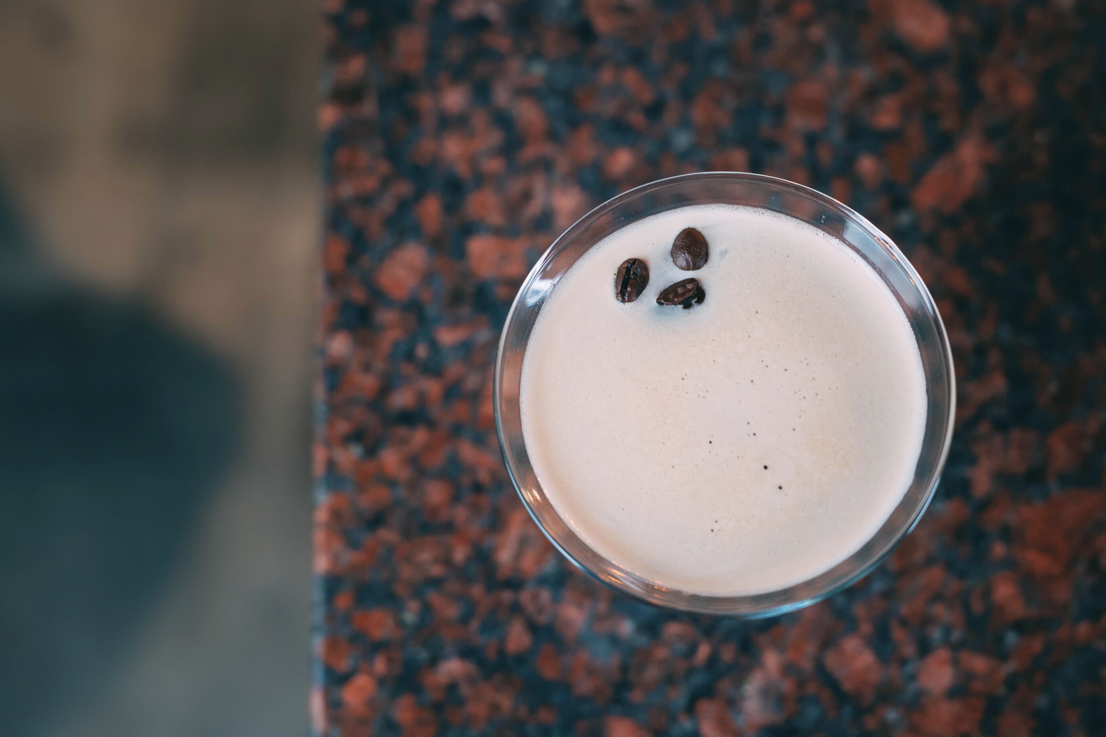 Un vaso con una bebida cremosa y dos granos de café sobre una encimera moteada.