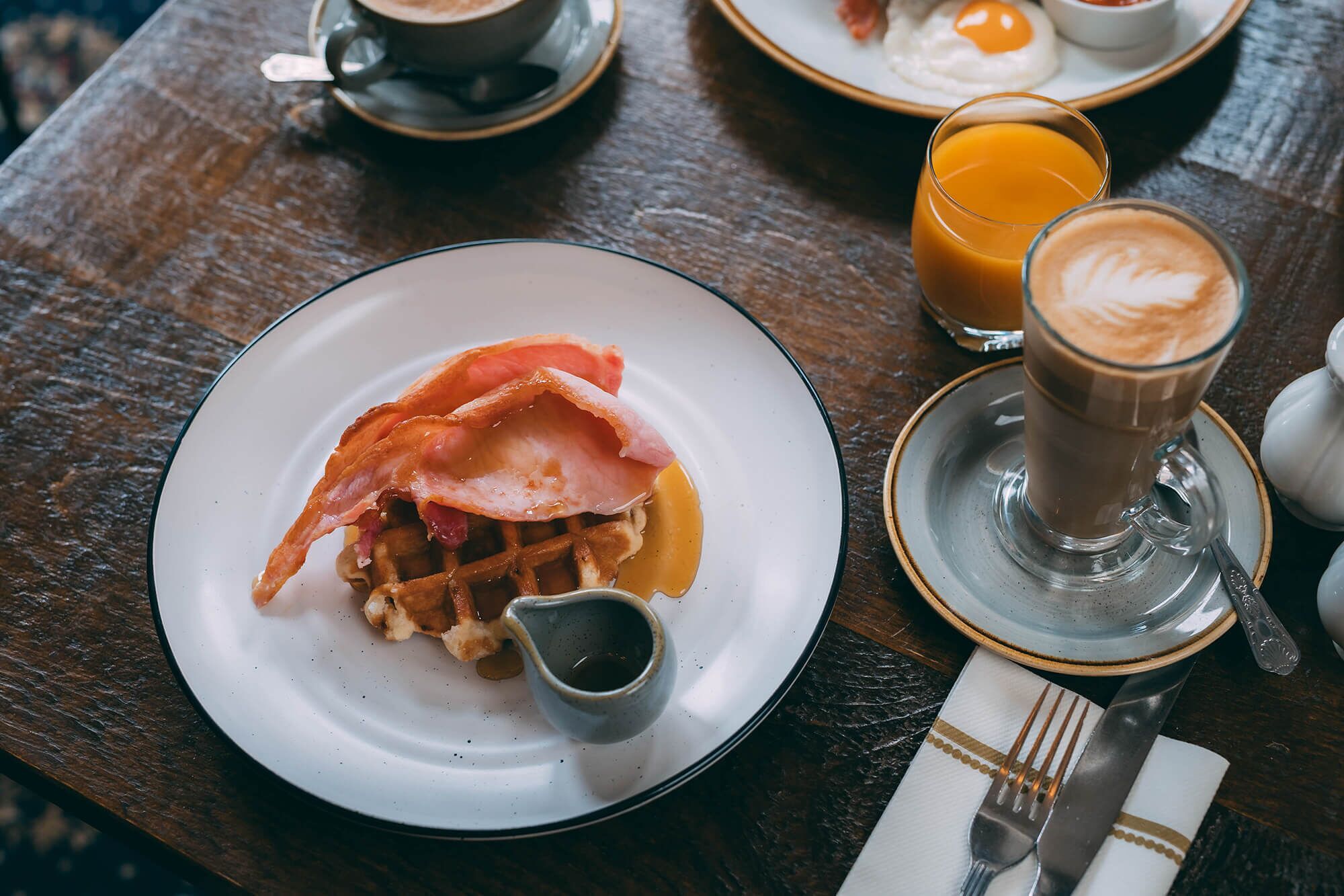 A breakfast setting with waffles, bacon, syrup, coffee, and orange juice on a wooden table.