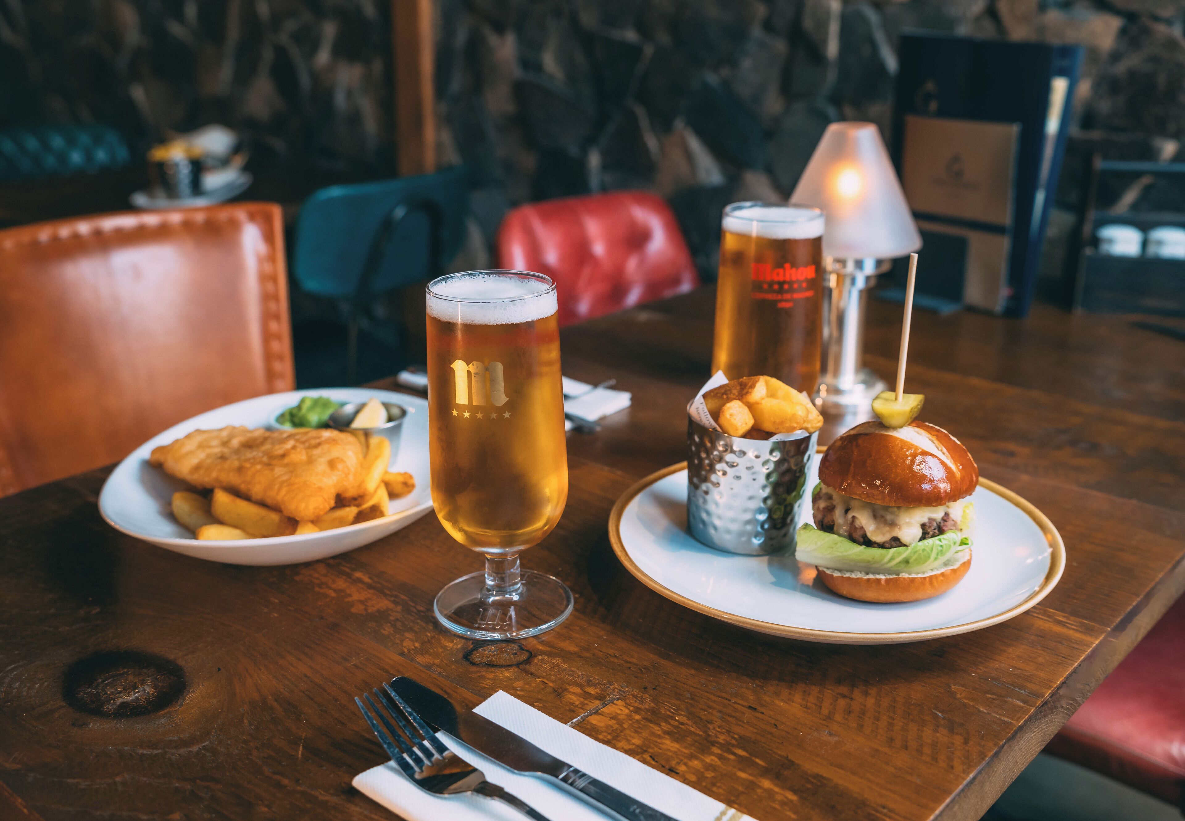 Una mesa de comedor con una comida que incluye pescado con papas fritas, una hamburguesa y dos vasos de cerveza.