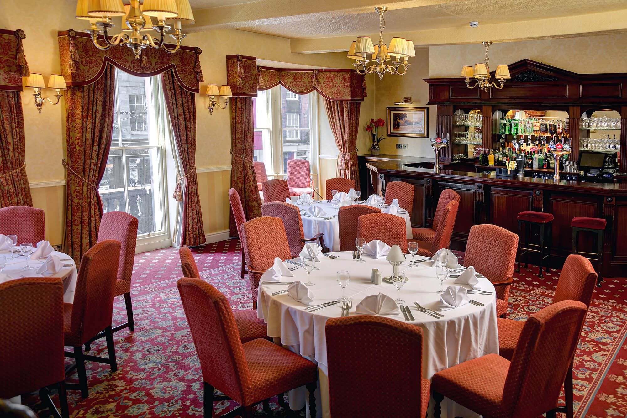 A restaurant with round tables set for dining, red upholstered chairs, a bar area with a variety of bottles, and a view through large windows.