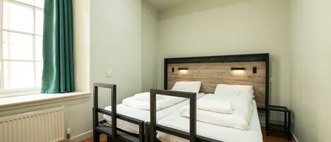 A bedroom with a wooden headboard, white bedding, a window with curtains, and a radiator.