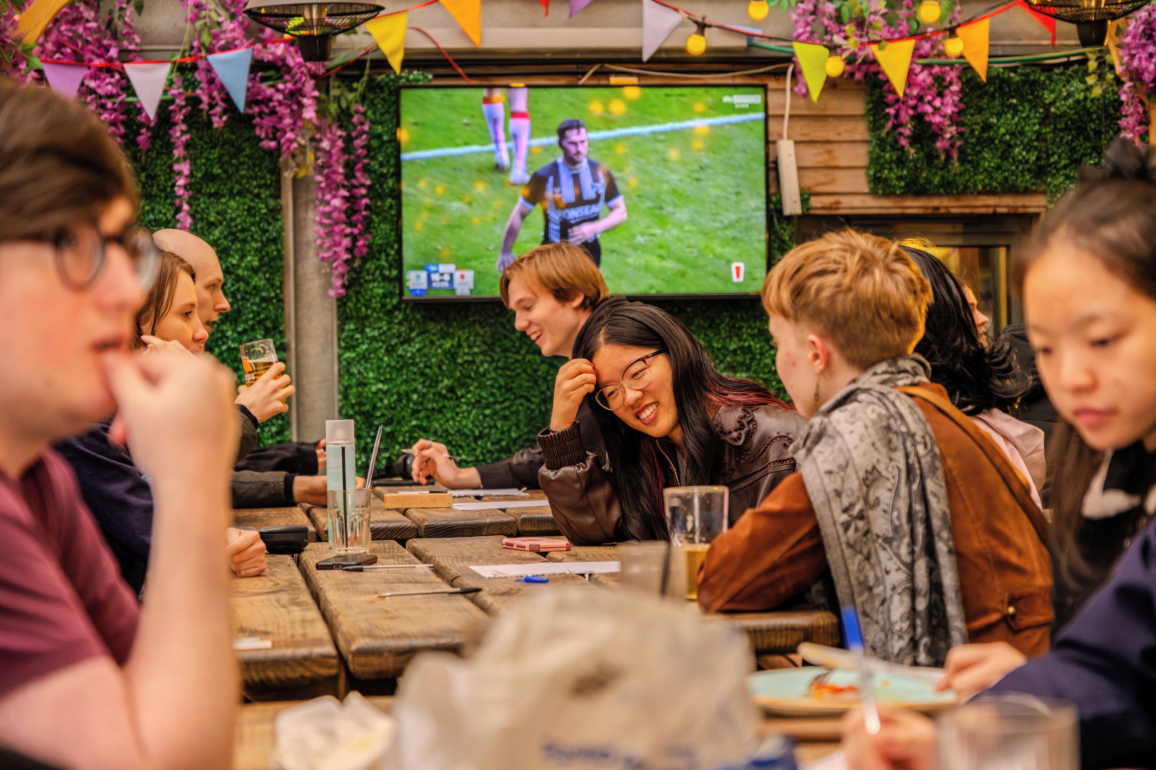 A group of people seated at a wooden table with a television screen displaying a sports event.