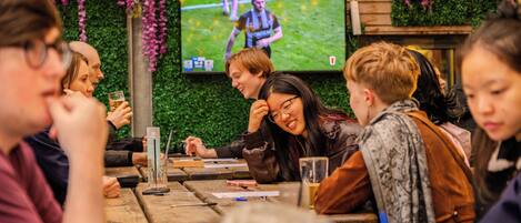 A group of people seated at a wooden table with a television screen displaying a sports event.