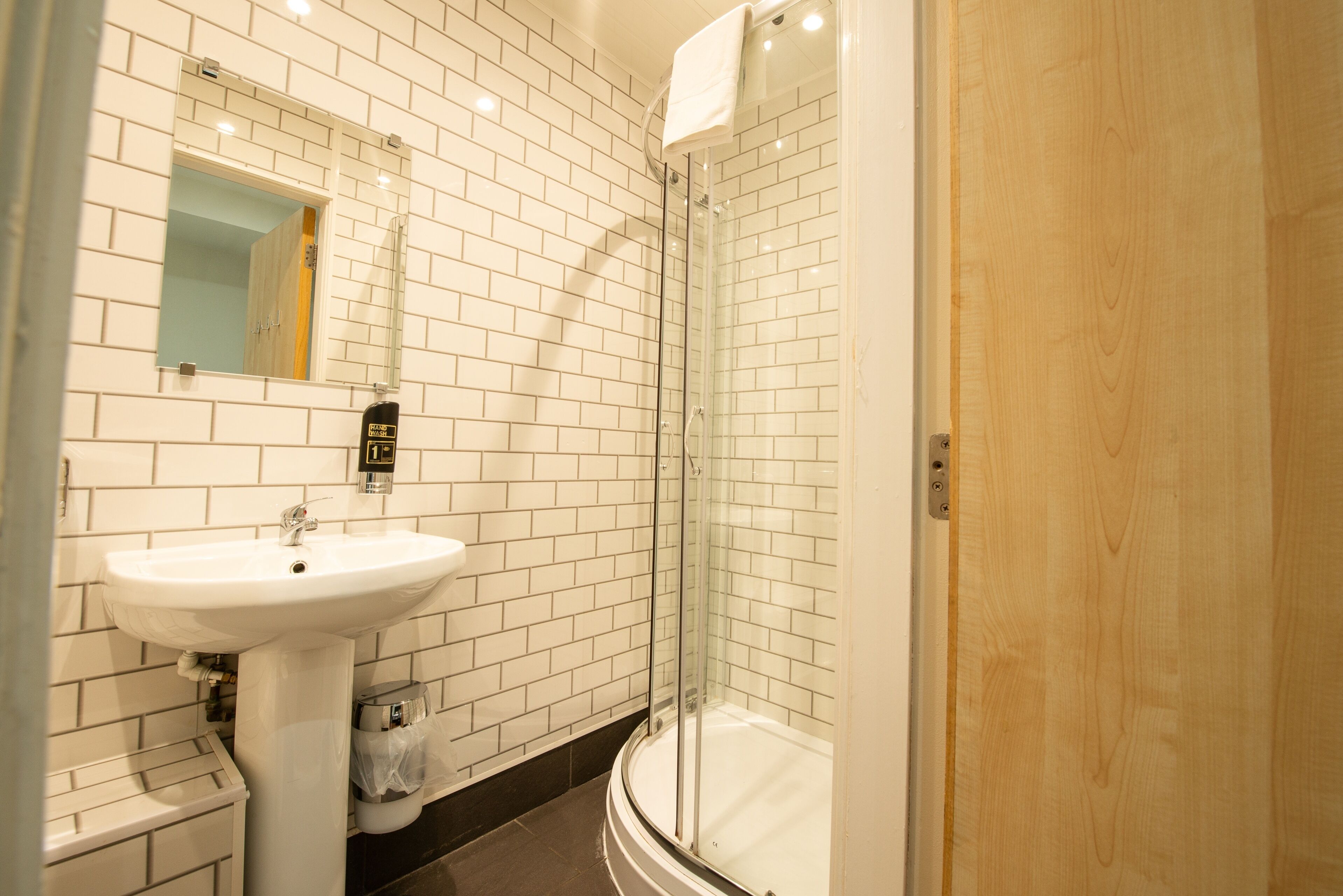 Un baño con pared de azulejos blancos, un lavamanos blanco y una ducha con mampara de vidrio.