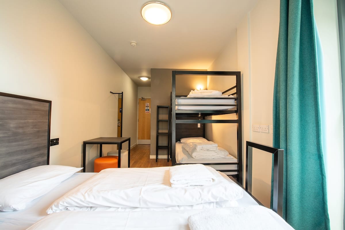 A hotel room with a bunk bed, a single bed, a small table, and a door leading to a hallway.